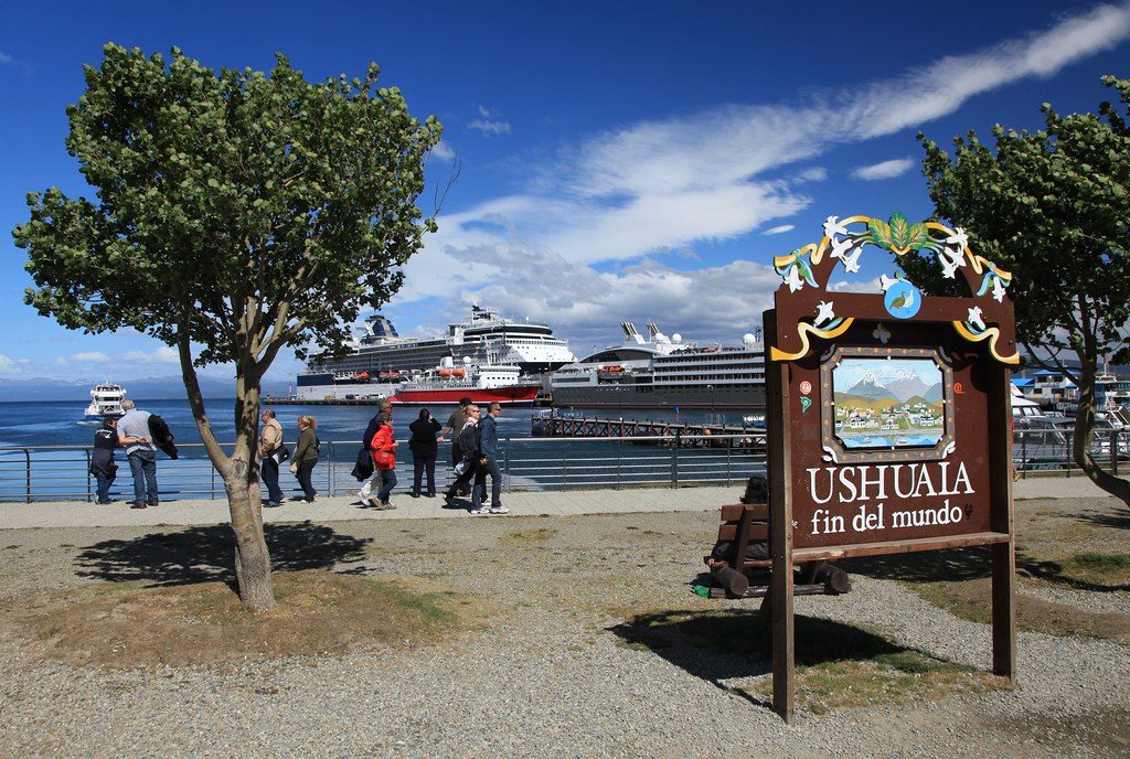 Туристы у знака Ushuaia Fin del Mundo и круизный лайнер в порту, городская экскурсия