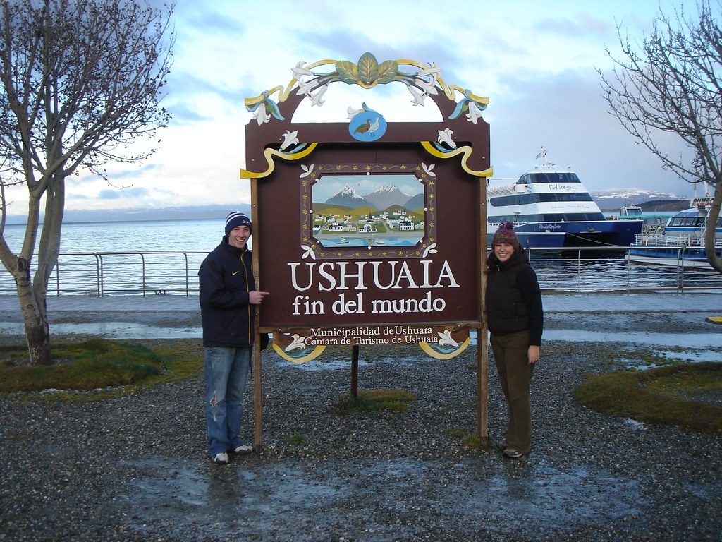 Туристы у деревянного знака Ushuaia Fin del Mundo в порту с лодками